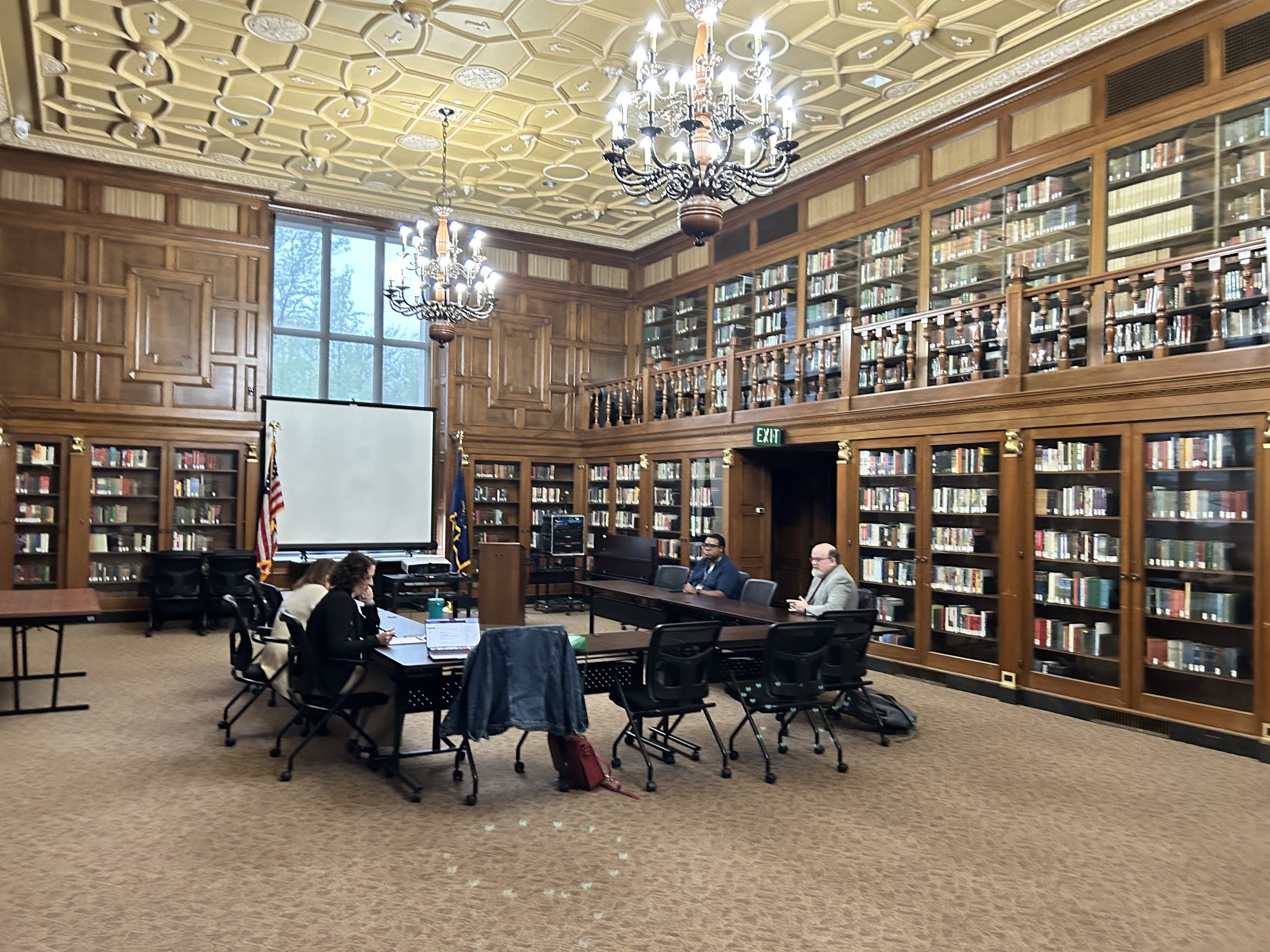 The Indiana Authors Room at the Indiana State Library has wooden book shelves lining the walls filled with books behind glass doors. In the center of the room is a u-shaped table setup with chairs in front of a projection screen.