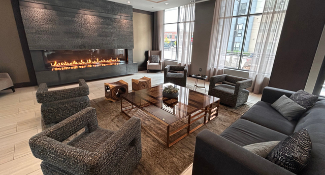 A cozy seating area in the lobby, with a couch and a variety of chairs. One wall nearby hosts a fireplace; the other has floor to ceiling windows letting in lots of natural light.