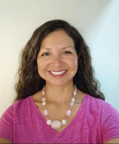 A photo of Maureen, a woman with light brown skin, freckles, wavy brown hair, and a big smile. She is wearing a bright pink shirt and a necklace with large, light pink beads.