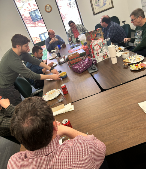 An angled view of the staff gathered around the conference table as they finish of their lunch. A pile of gifts waits temptingly in the middle.