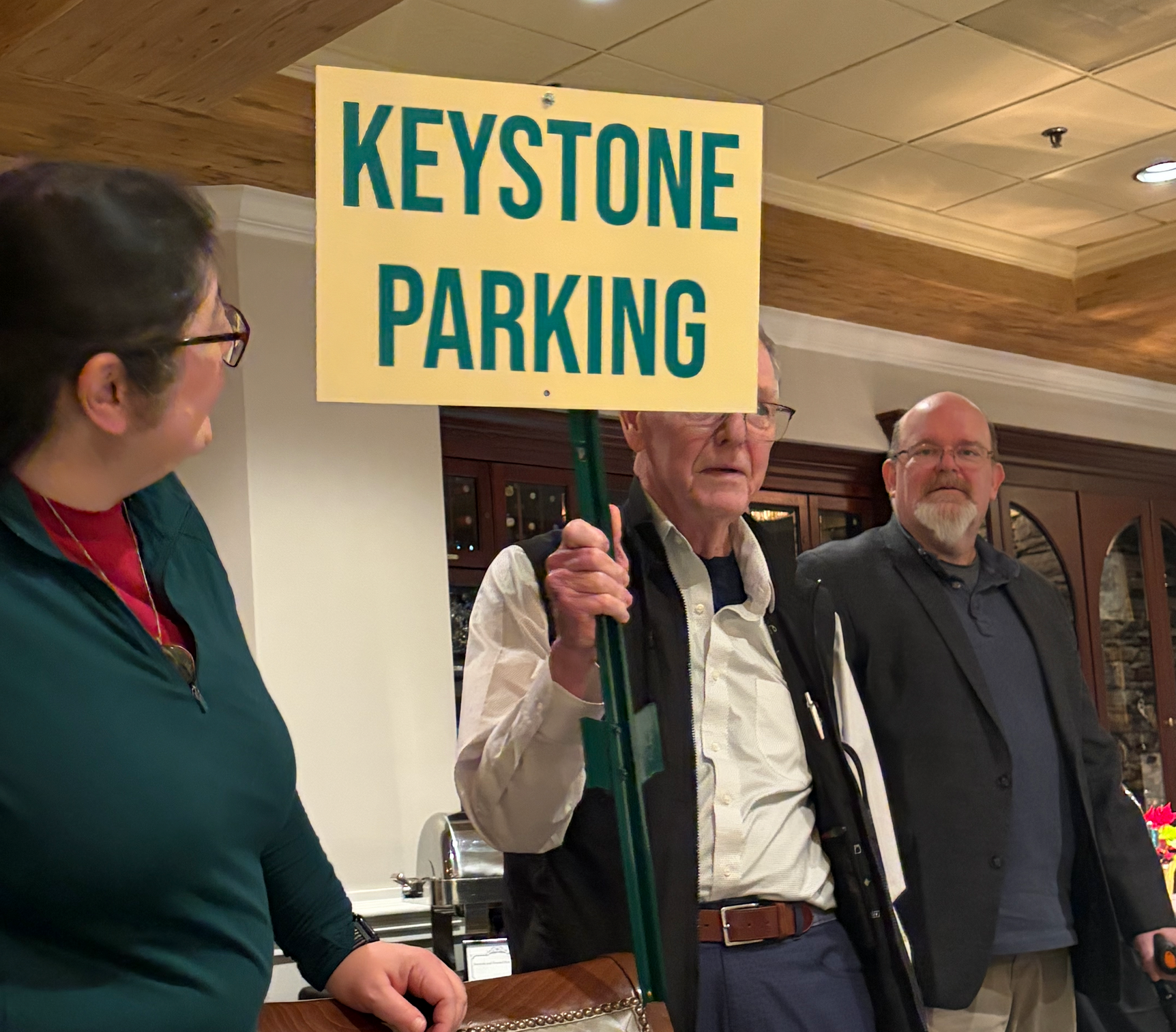 Mitake and James look on as Dave unveils a "Keystone Parking" sign, printed big and bright, although it still needs a second bolt affixing it to its signpost.