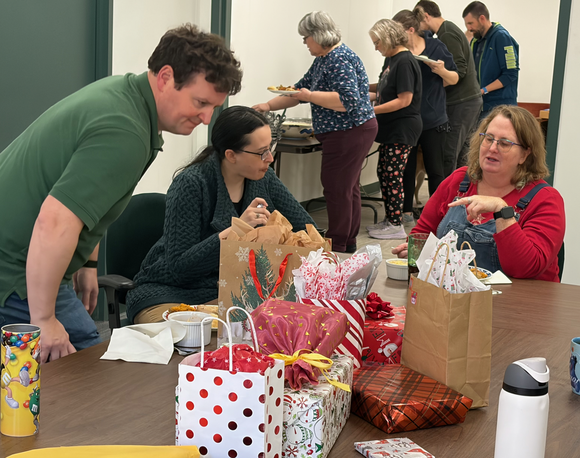 John, Katy, and Marion settling in at a conference table with gift bags and wrapped presents clustered in the middle, while Nancy, Tracey, George, and Lee go through the buffet line behind them.