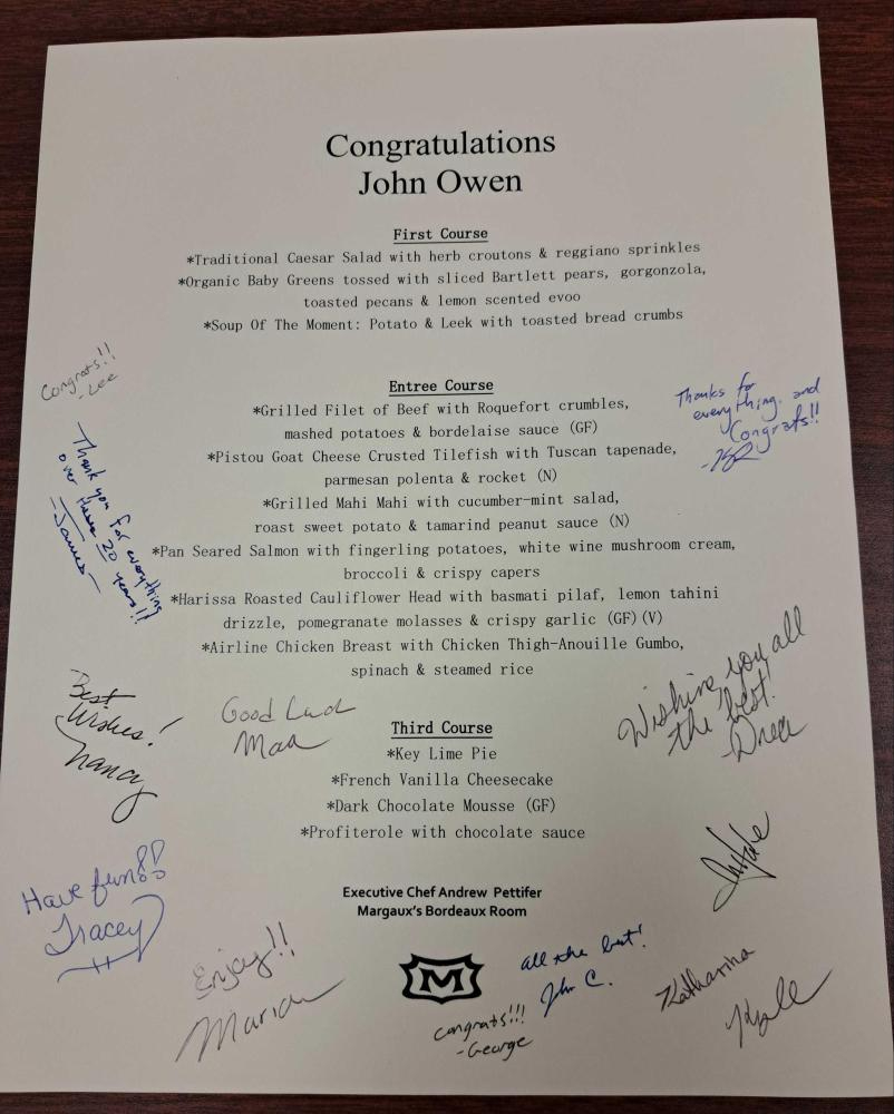 A photo of the menu, which everyone has signed for John. At the top, it reads Congratulations John Owen, then it lists the options for each course with Margeaux's logo at the bottom. The Keystone staff have written their congrats and well-wishes around the menu text in blue or black ink.