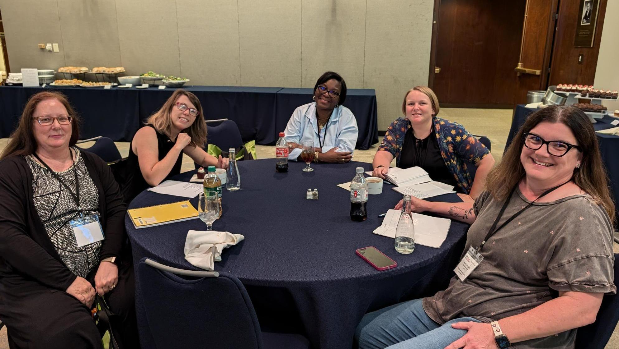 Five female KLAS Users sit at a round table covered in a black tablecloth. They are all facing the camera and smiling. A variety of pens, notebooks, drinks, and other items are on the table in front of them.