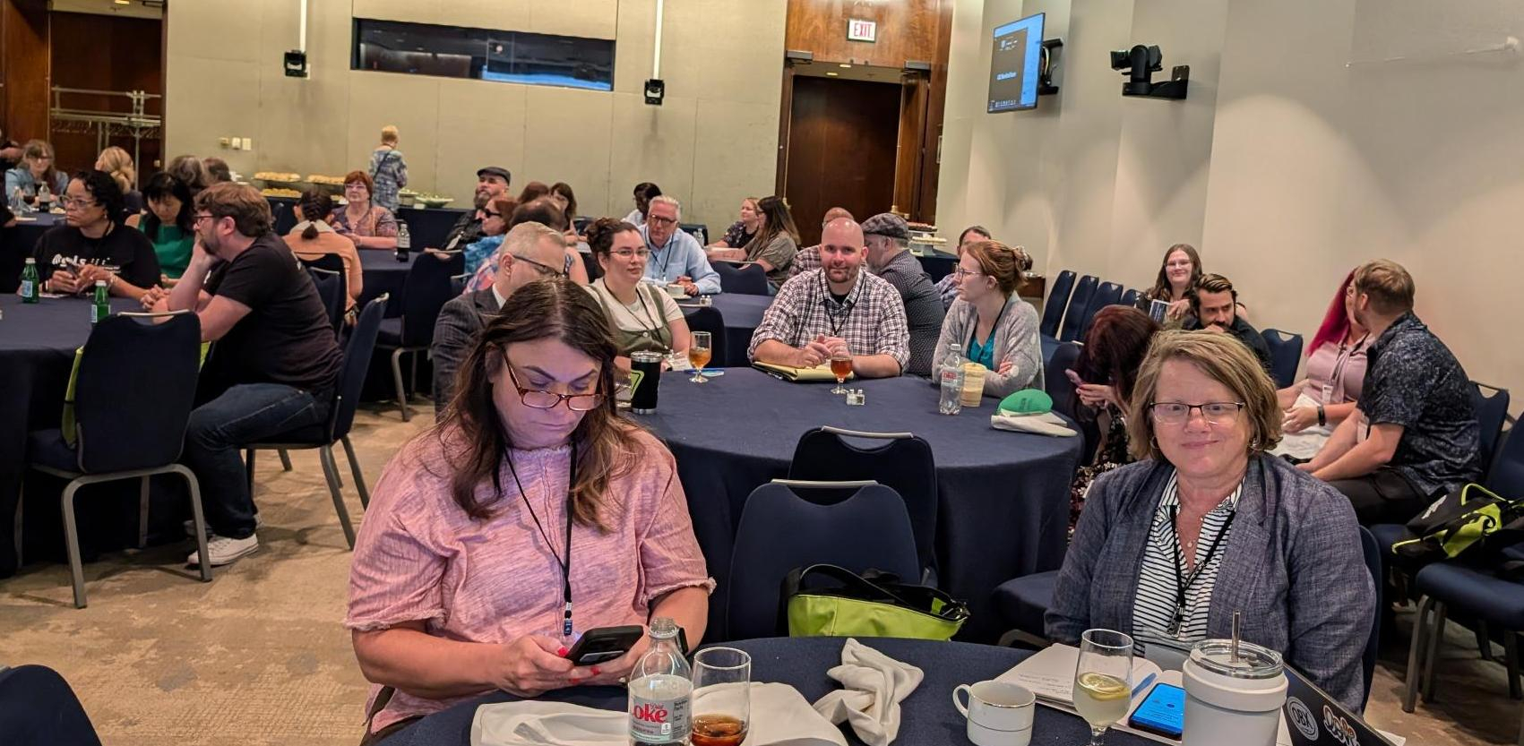 A photo shows a room with NLS conference attendees seated at multiple round tables draped with black tablecloths. In the foreground, are two female attendees. One is looking at the camera and smiling and the other is looking at her phone.