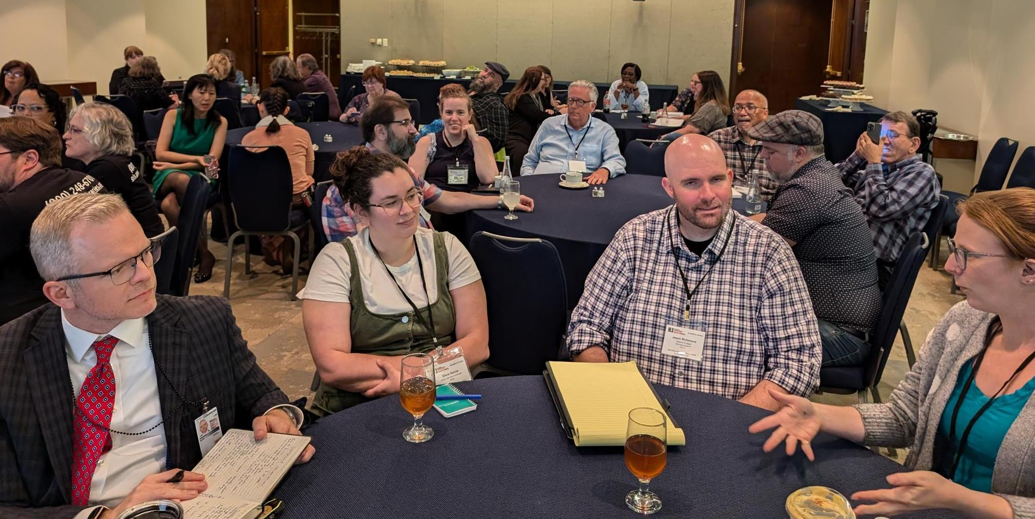 A group shot of NLS attendees seated at round tables with black tablecloths. Four people (two women and two men) are the table in the foreground are having in a lively discussion.