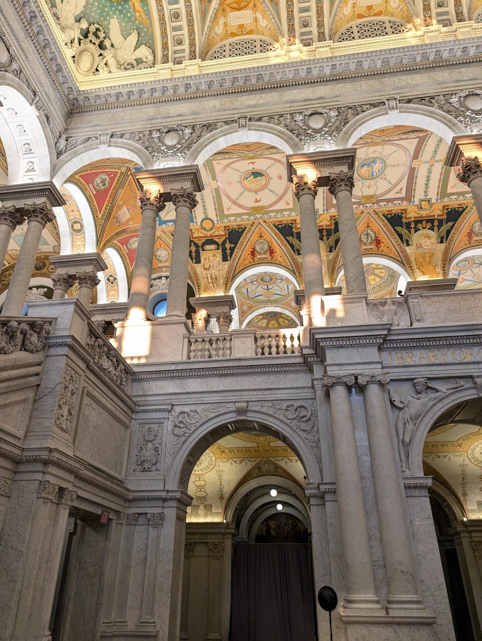 A colorful, majestic interior shot of ornate architecture. Huge arches support a second floor balcony, which itself has elegant columns to support the vaulted ceiling. The ceilings are entirely painted, and the stone walls and supports are carved in a classical style.