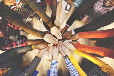 A wide-variety of people stand in a circle and put their hands in to show teamwork.