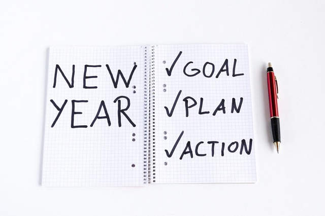 A spiral notebook lays open next to a pen with "New Year" written on the left page and checked off items on the right including: Goal, Plan and Action.